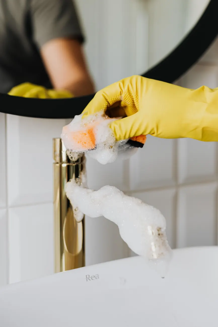 Gloved hand polishing a brass faucet with a soapy sponge